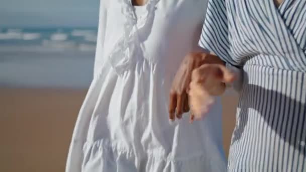 日光の中で海の海岸を歩いてカップルの手を閉じます 海の岸で腕をつないで行くデートを楽しむガールフレンド 休暇中にリラックスして話している認識されていないLgbtのパートナー 多民族のLgbt関係 — ストック動画