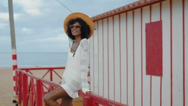 在阴郁的一天 微笑的女人在海滩上休息 快乐的非洲裔美国人喜欢在海滨别墅度假 戴着草帽太阳镜 身材苗条的漂亮女孩穿着白色的泳衣放松 Idyllic海洋周末概念 — 图库视频影像