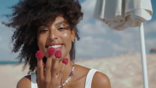 美しい女の子の指を閉じてラズベリーを食べる 夏の日差しの中で健康的な軽食を楽しむ笑顔の女性の残りのビーチ ベリーと遊ぶ幸せな魅力的なアフリカ系アメリカ人 有機おいしい食べ物 — ストック動画