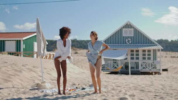 ビーチの女の子はビーチハウスで日光浴をオフに服を取る ビキニのスリム女性は海岸で夏休みを過ごしています 休日を楽しむ魅力的な多民族Lgbtカップルパートナー — ストック動画