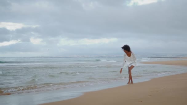 一个性感的女人独自在海洋海岸线上行走 快乐的无忧无虑的女孩穿着白色的衣服触摸沙滩捕捉海浪 放宽梦幻般的非洲裔美国人享受岛屿周末 和平假日度假概念 — 图库视频影像