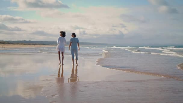 夏の日差しの中でレズビアンカップルウォーキングビーチ 2人の親友が周りを見て海の海岸を休んでいる リラックスした女性多民族の愛好家は海岸で休暇を過ごす 観光休暇リゾートのコンセプト — ストック動画