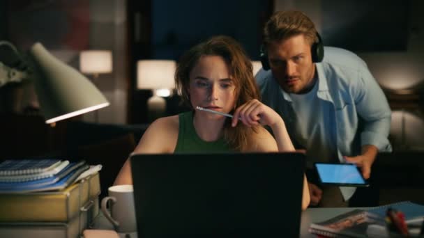 夜のフラットクローズアップで集中的な女の子の読書装置 コンピュータを見ている思慮深い女性は夫を踊ることに注意を払っていません フリーランスの女性がノートパソコンを閲覧 肩から画面を見ている男 — ストック動画