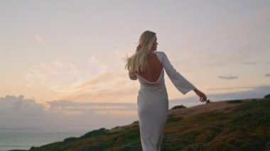 Akşamları rahatça yürüyen bir kadın. Uçan saçlı kız gün batımı vadisinin doğasında poz veriyor. Doğal güzellik modeli, kamera görünümlü, saç stiline dokunuyor. Sarışın kadın bulutları döndürüyor.