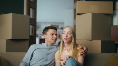 Kiralık eve taşınan pozitif genç çift rahat koltuğa yakın oturuyor. Mutlu aile, modern oturma odasında kutu dolu kutular içinde dinleniyor. Neşeli çift yeni bir daireyi tartışıyor..