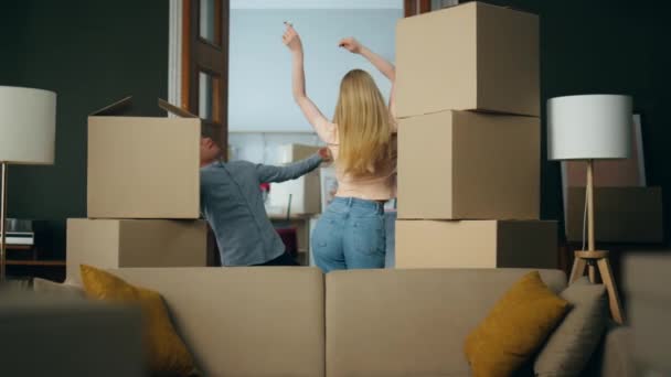 新的快乐的房主们在纸盒包装的盒子之间欢快地跳舞庆祝他们自己的房子 年轻而积极的家庭因为搬进新公寓而高兴地在一起鬼混 迁移概念 — 图库视频影像
