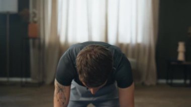 Çekici genç sporcu eğitimini evde tek başına yakından izle. Diz çöküp spor yapan kaslı adam kameraya baş kaldırıyor. Dövmeli sağlıklı, ciddi bir adam. Fiziksel sağlığını önemsiyor.