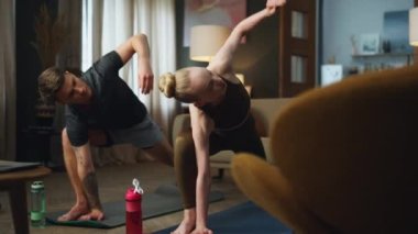 Evde yoga pozu veren genç sporcu çift. Esnek adam, içeride sarışın bir kadınla bacaklarını esneterek üçgen pozisyonunu uyguluyor. Milenyum ailesi vücut sağlığını önemsiyor.