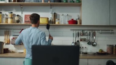Neşeli adam rahat mutfakta tek başına yemek pişirerek dans ediyor. Çekici genç adam, sabahları lezzetli bir kahvaltı hazırlarken modern laptopta müzik açıyor. Mutlu aşçı evde bilgisayar kullanıyor..