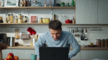 Dizüstü bilgisayarla müzik açan mutlu adam mutfak masasını yerleştirdi. Rahat evde mavi tişörtlü neşeli adam dans ediyor. Genç tatmin olmuş serbest çalışan apartmanda uzaktan çalışmaktan zevk alıyor..