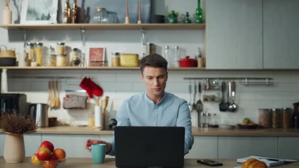 若いリモートワーカーは自宅からノートパソコンでコーヒーを飲んでいます キーボード上のコンピュータ画面の書き込みテキストを探している事業者に焦点を当て 魅力的な男感じリラックスした仕事をオンラインで現代的なキッチン — ストック動画