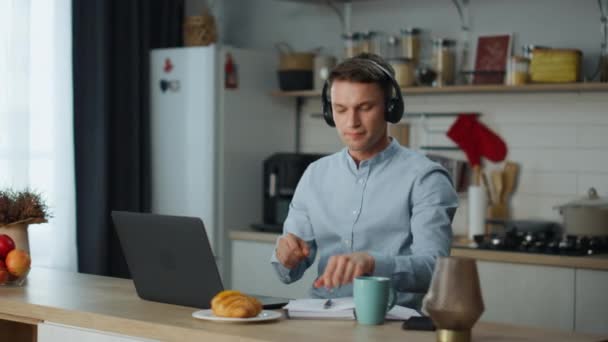 リモートワーカーは オンラインネットワーク接続でノートパソコンを使用して自宅で働くヘッドフォンから音楽を聞きます キッチンに座ってフリーランスの仕事を楽しんで踊る積極的な男 無線ヘッドセットを身に着けてる — ストック動画