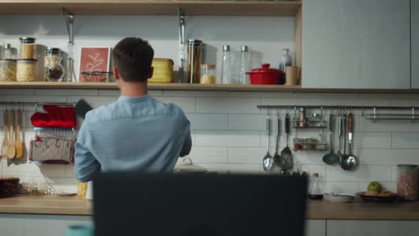 一人で朝食の準備をするへらで台所で踊る幸せな若い男 木製のテーブルの上に立ってノートパソコンでビデオ通話に答える魅力的な陽気な男 現代のアパートでハンサムな男の料理 — ストック動画