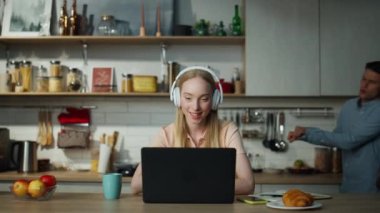 Evde kablosuz kulaklık takan güzel bir kadın internet yöneticisine internet üzerinden danışmanlık yapıyor. Arka planda dans eden neşeli genç adam. Çekici kız yakınlaştırma konferansında dizüstü bilgisayarla konuşuyor..