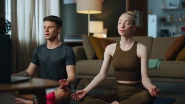 Genç ve rahat bir çift evde meditasyon yapıyor. Sakin bir çift nilüfer, laptopta ruhani dersleri dinlerken kapalı gözlerle spor minderleri üzerinde poz veriyor. Çekici kadın bir erkekle yoga yapıyor..