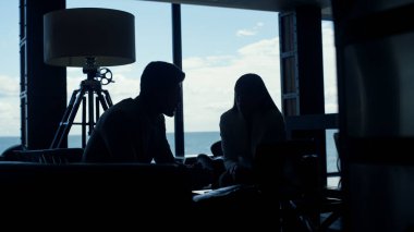 Siluet, okyanus manzaralı lüks ofiste konuşan rahat bir çift. Panorama pencere lobisinde çalışan tanınmamış odaklanmış insanlar. Aşk çifti özel karanlık iç atmosferde flört ediyorlar. 