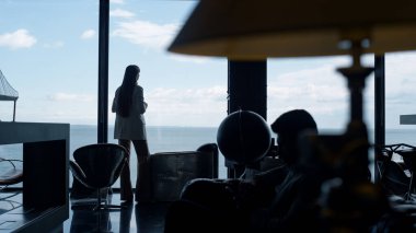 Deniz manzaralı ofiste rahatlamış iş kadını silueti. Tanınmayan lider tek başına panorama penceresini izliyor. Şirket çalışanı toplantıdan sonra dinleniyor. Düşünceli üst düzey yönetici arka plan çekimi