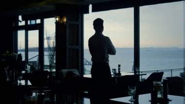 Pensive man silhouette standing restaurant hall alone. Attractive guy enjoying beautiful ocean view in panoramic lounge bar. Thoughtful boss looking window in dark interior. Business success concept. clipart