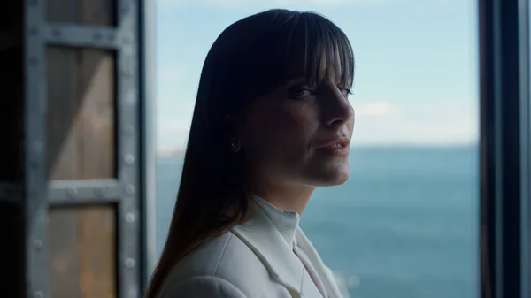 Closeup businesswoman smiling ocean window view. Corporate professional turning head to camera. Woman director face expression. Confident boss enjoying journey indoors. Ambitious person concept