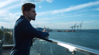 Kendine güvenen işadamı, sabah güneşinde deniz limanı vinçleri manzaralı balkonda dinleniyor. Ciddi bir işadamı cam kafe terasında dinleniyor. Çekici adam profesyonel müşteri toplantısı.