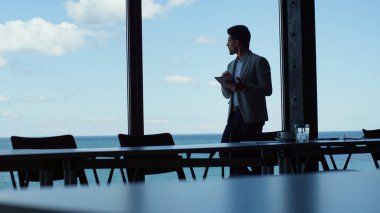 Focused entrepreneur using tablet in sea view dark office building. Confident businessman browsing digital device by panoramic window. Professional watching marine landscape researching data on pad  clipart