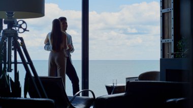 People silhouettes in love embracing sea view luxury hotel. Unknown romantic couple dating against ocean panoramic window. Rich man hugging wife waist tenderly at marina landscape room. Lovers concept