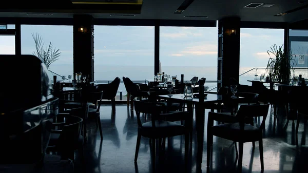 Spacious panoramic restaurant hall interior in evening dusk background. Stylish dark hotel rooftop cafe dining area. Magnificent horizon line sea view in modern sky bar. Dusk business lunch concept.