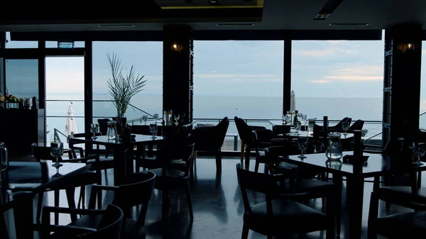 Dark interior design in modern panoramic hotel bar. Scenic evening ocean view in luxury loft lounge cafe. Unknown man waiter silhouette walking stylish restaurant hall in dusks.