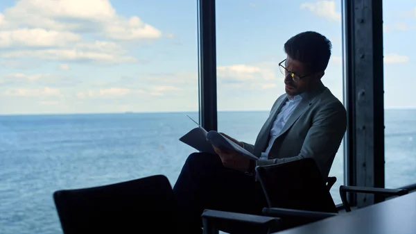 Businessman reading contract papers at sea view. Thinking manager analyzing data looking panorama window. Focused man lawyer checking finance documents corporate report. Male professional at work.