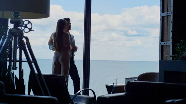Sensual couple enjoying ocean view at hotel. Newlyweds silhouettes cuddling at panoramic window place vacation. Unknown man hugging woman waist. Lady and guy embracing together. Honeymoon concept