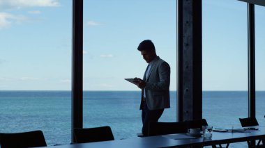 Thinking businessman using tablet in panoramic window office. Executive standing working online at sea view dark hall. Successful entrepreneur searching device. Ceo dreaming career opportunities