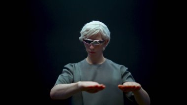 VR elemanı arttırılmış gerçekliği yakından araştırıyor. Futuristik gözlüklerle odaklanmış oyuncu karanlık arka planda metaevren simülasyonu yaşıyor. Siber sanal nesnelere ışık tutan şık sarışın adam..