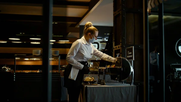 Masked hostess working dark restaurant interior alone. Elegant waitress setting breakfast food on cafe buffet table. Beautiful hotel bar employee serving lunch. Protective measures at covid19 concept