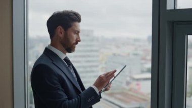 Ofis panoramik penceresinde duran tablette çalışan kendine güvenen sakallı bir iş adamı. Sekme bilgisayarında yazan başarılı yöneticinin portresi şirket perspektifleri üzerinde düşünen geliştirme başarısı.