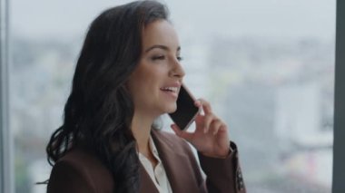 Gülümseyen kadın ofis müdürü pencerenin önünde telefonla konuşuyor. Modern akıllı telefon kullanarak müşterilerle konuşan çekici, zarif bir iş danışmanı. Mutlu işçi danışmanı telefonda..