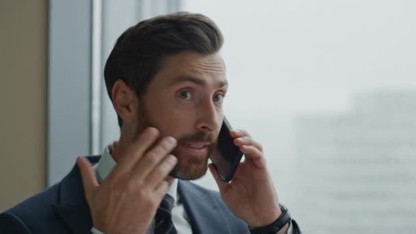 オフィスの窓で感情的に立って失望したビジネスマンの話電話を閉じる 怒っている髭のマネージャーはスマートフォンで不快な会話をして叫んでいます 緊張した男がストレスを感じる — ストック動画