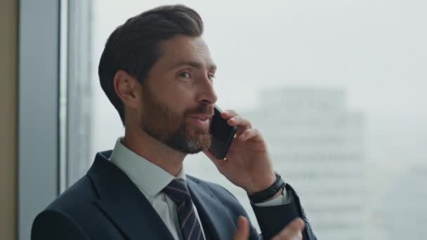 エレガントなスーツを着てオフィスの窓に立って携帯電話で話すハンサムな自信のある起業家を閉じます 仕事の合間に電話でのコミュニケーションを楽しんでいるビジネスマンのCeoを笑顔 商談会 — ストック動画