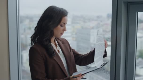 優雅なスーツ姿でオフィスの窓辺に立って契約書を読む若い成功した女上司 魅力的なビジネスウーマンマネージャーの肖像会社の財務結果と幸せな表情の論文を感じます — ストック動画