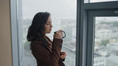 Ofis panoramik penceresinde kahve içen kendine güvenen çekici bir iş kadını. Şehir manzarası çok yakın. Zarif esmer kadın müdür. Sıcak içeceklerin keyfini çıkar. İş molası.