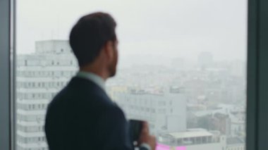 Gülümseyen CEO sıcak çay içiyor ofis penceresine bakıyor ve başarıyı yakından hissediyor. Çekici sakallı işadamı, kârlı bir ortaklık anlaşması imzaladıktan sonra kahve molasının keyfini çıkarıyor..