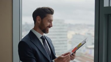 Gülümseyen CEO ofis penceresinin önünde duran mali belgelerden memnun kaldı. Mutlu başarılı iş adamı satış istatistiklerini okuyor kârlı yatırım projesi muhasebe raporu.