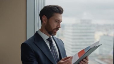 Finansal analist ofis penceresinde duran iş belgelerini inceliyor ve iş sonuçlarını yakından düşünüyor. Turta grafiğinde satış istatistiklerini okuyan kendine güvenen işadamı, şirketin verimlilik verilerini analiz ediyor..