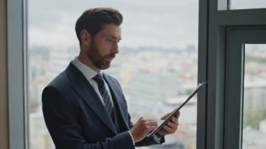 Ciddi yorgun işadamı ofis penceresinde duran şirket belgelerini kontrol ediyor. Yönetim kurulu başkanı, mali evrakları analiz eden belgeleri okuyor. İş için endişelenen orta yaşlı sakallı adam..