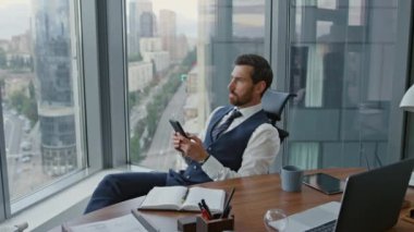 Ciddi bir girişimci elinde akıllı telefon, panoramik pencerenin yanında oturuyor. Modern iş yerinde cep telefonu kullanan odaklanmış bir iş adamı. Düşünceli şirket çalışanı cep telefonundan mesaj yolluyor.. 