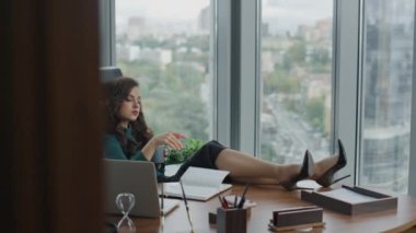 Kendine güvenen çekici CEO kadın ofis masasında oturuyor ve modern dizüstü bilgisayarla kariyerini düşünüyor. Başarılı esmer menajer, tahta masaya bacaklarını koyuyor ve iş fikirleri yaratıyor. İş kadını dinleniyor
