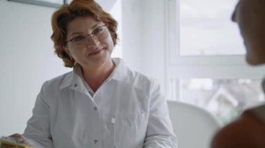 Orta yaşlı, gözlüklü kadın doktor estetik cerrahi cilt bakımı için hastaya danışmanlık yapıyor. Tıp kürklü profesyonel hekim masaya oturup tedaviyi yakından tartışıyor..