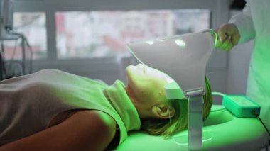 Kadın kozmetoloji kliniğinde yüz yenileme terapisi görmüş. Bilinmeyen estetik uzmanı ışık maskesini ayarlayan fototerapi cihazını kontrol ediyor. Güzellik salonunda yüz bakımı.