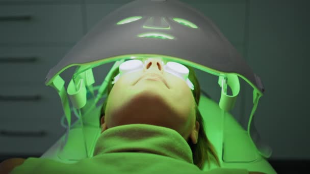 在美容外科诊所 松驰的妇女躺在头戴浅色抗衰老面罩下进行面部肌肤更新 戴眼镜的女士正在做美容美发程序 非侵入式光疗 — 图库视频影像