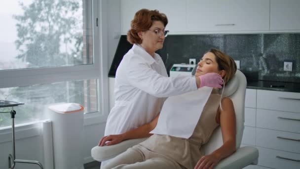 美容の手順に女性のクライアントを準備認定美容師皮膚科医 医者は保護ナプキンをつけてる 魅力的な女性は相談美容クリニックで白いモダンな椅子に座って — ストック動画
