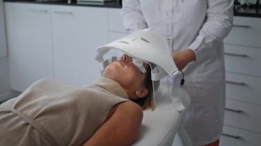 Bilinmeyen kozmetik uzmanı fototerapi için özel öncü maskeyi çalıştırıyor. Kadın müşteri kırışıklıkları düzeltmek için modern fototerapi ekipmanları altında yatıyor. Güzellik kliniği kavramı.